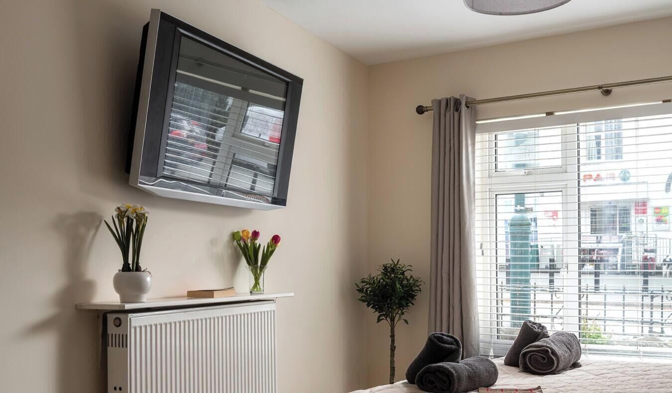 A hotel room with a bed, a radiator, a TV mounted on the wall, and a window with curtains.
