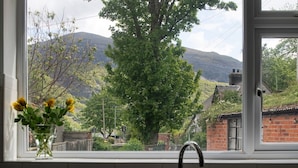 Une fenêtre donnant sur une montagne, un arbre et un bâtiment en briques.