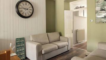 Ein Wohnzimmer mit beiger Couch, einer Wanduhr, einem motivierenden Spruch und einer Küchenzeile.