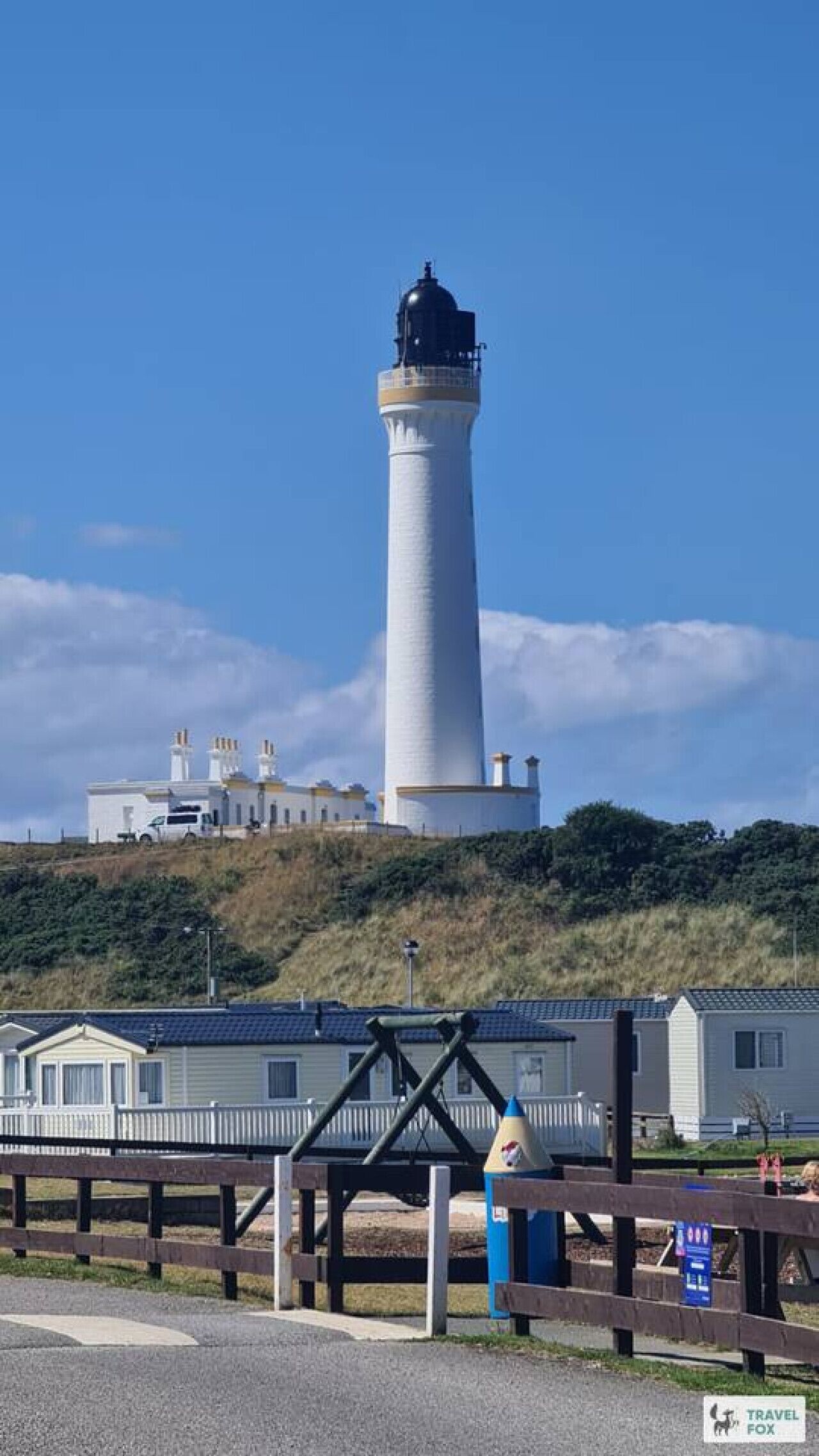 Un phare avec une tour blanche et un sommet noir, un bâtiment blanc doté de plusieurs cheminées, et une rangée de maisons mobiles.