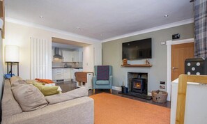 A living room with a sofa, a chair, a fireplace, a television, and a kitchen in the background. - Daisy Cottage - Hot tub and fire pit! (Aberdeenshire)