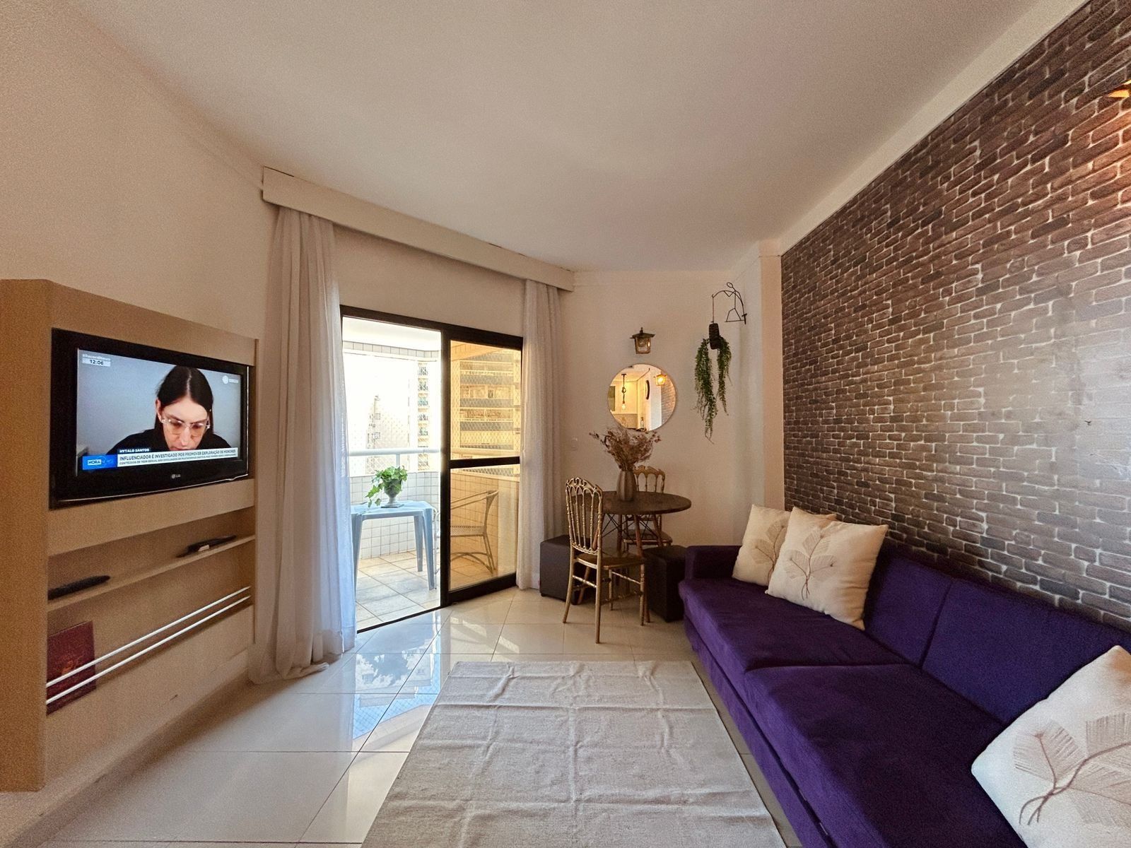 A living room with a TV, a purple sofa, a dining table with chairs, and a brick wall.