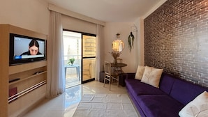 Una sala de estar con televisión, un sofá morado, una mesa de comedor con sillas y una pared de ladrillo.