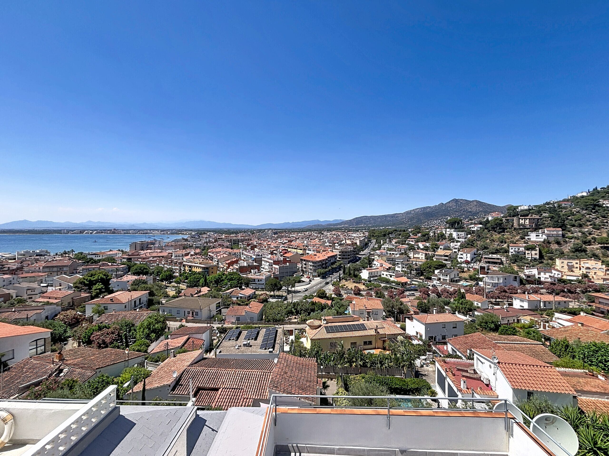 A coastal town with terracotta-roofed buildings, a clear blue sky, and a view of the sea and distant mountains.