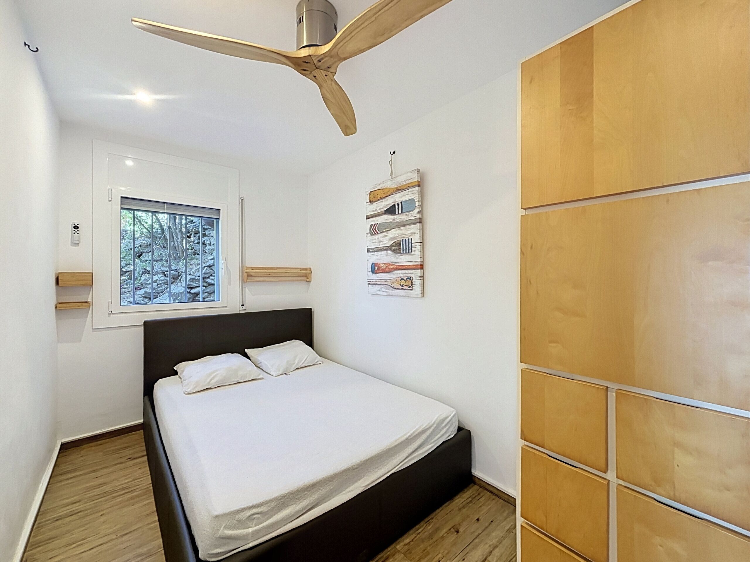 Un dormitorio con cama, ventilador de techo, ventana con vistas y armario de madera.