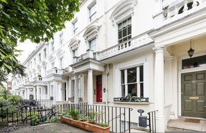 A row of white townhouses with balconies, a red door, and a black metal fence. - The Hummingbird 1 Bedroom by Kensington Gardens (London)