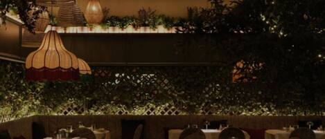 Um restaurante com iluminação suave, mesas e cadeiras, luminárias pendentes e uma parede coberta por vegetação.