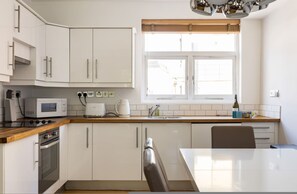 Eine moderne Küche mit weißen Schränken, einer Holz Arbeitsplatte, einer Mikrowelle, einem Wasserkocher und einem Fenster mit Jalousien.