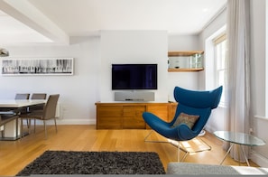 Ein modernes Wohnzimmer mit Fernseher, einem blauen Sessel, einem Esstisch und einem schwarzen Teppich.