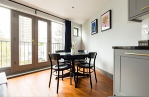 A dining area with a round table and chairs, a window with a view, and a kitchen area with cabinets. - Sunshine In My Mood 2 Bedroom Flat in Battersea (London)