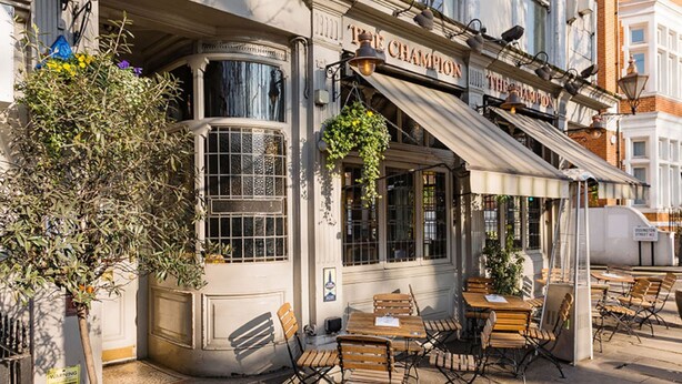An outdoor seating area with wooden tables and chairs, a hanging plant, and a building with a sign reading 'The Champion'. - Notting Haven - 2 BR Flat and Garden Notting Hill (London)