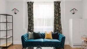 A living room with a teal sofa, a glass coffee table, a black shelving unit, and a white cabinet. - Notting Haven - 2 BR Flat and Garden Notting Hill (London)