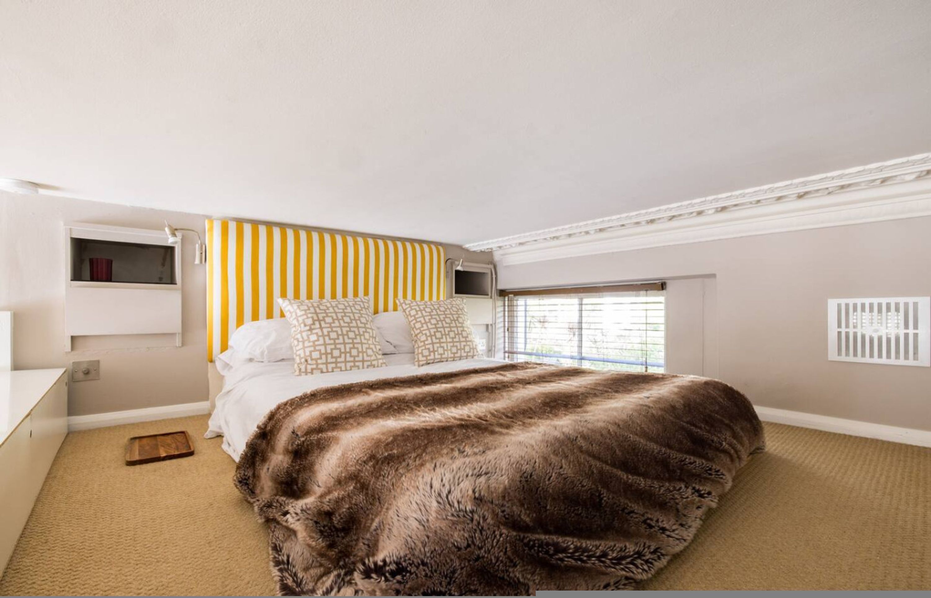 Quarto com cama grande, janela com persianas, mesa de cabeceira e cabeceira com listras verticais.