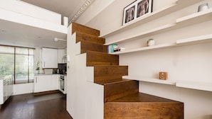 A modern kitchen with white cabinetry, a wooden staircase, and built-in shelves with decorative items. - Victorian Blush 1BR Near Portobello Notting Hill (London)