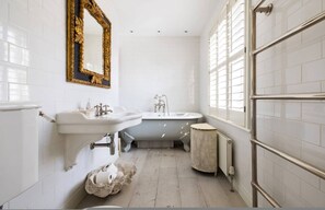 Une salle de bain aux murs recouverts de carreaux blancs, un grand miroir avec un cadre doré, une baignoire sur pieds et un petit tabouret rond.