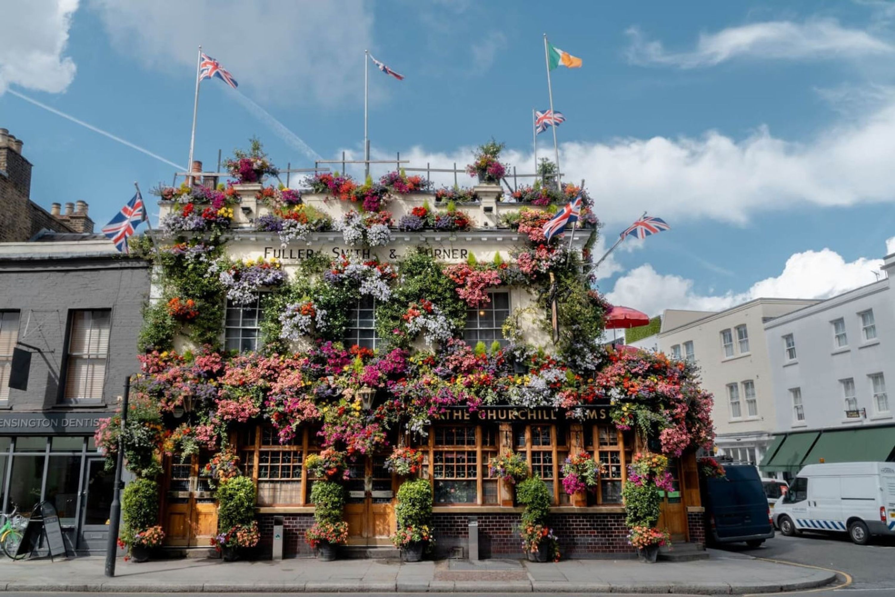 A building with a sign reading 'FULLER, SMITH & GARNER' and 'THE CHURCHILL HOUSE' with multiple flags and flower arrangements.