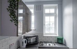Een kleine keuken met een inbouwoven, waterkoker, gasfornuis en een raam met zonwering.