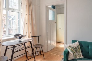 Een kamer met een raam, een tafel met stoelen, een groene bank en een deur naar een andere kamer.