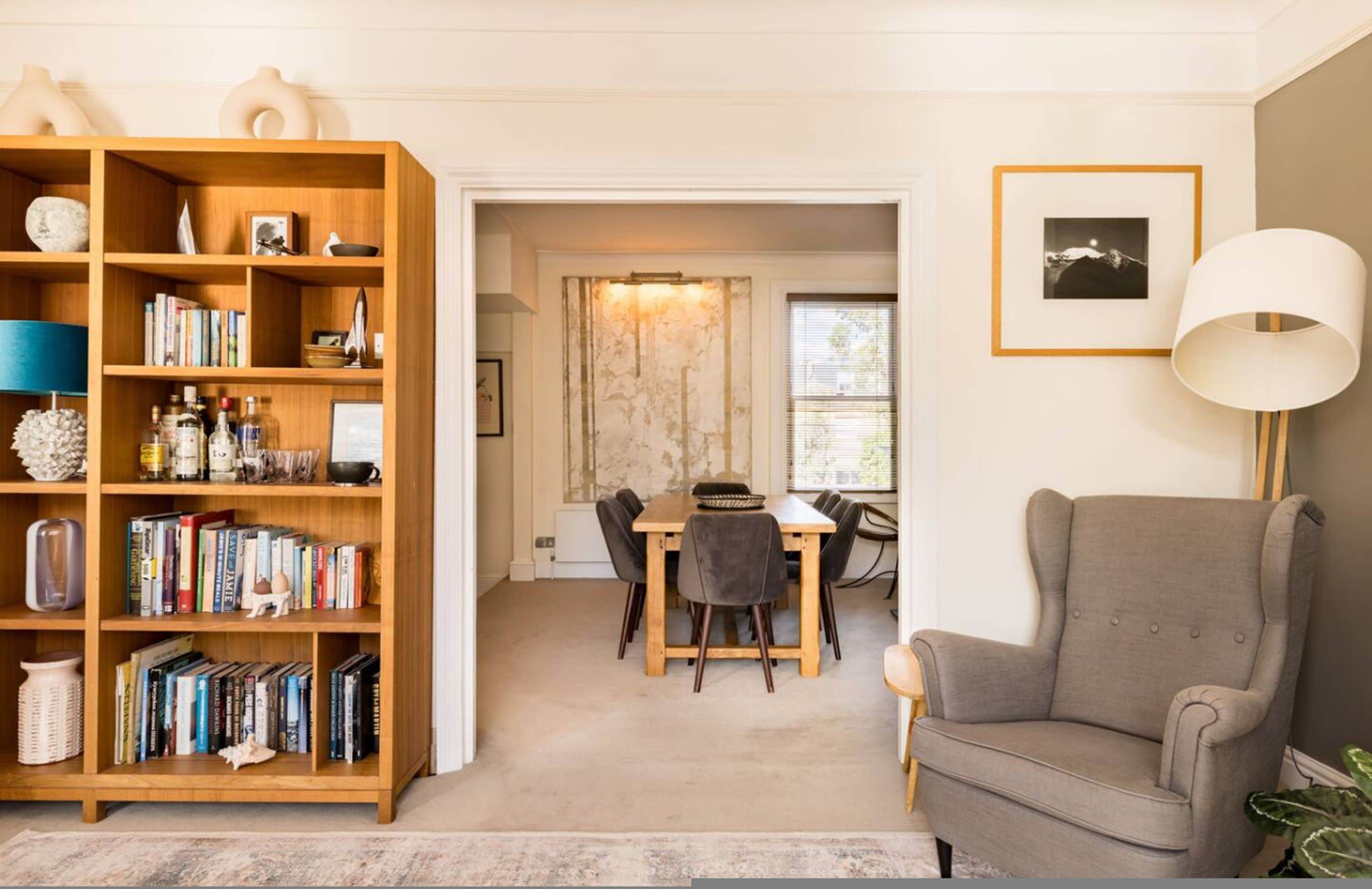 A living room with a bookshelf, a dining area with a table and chairs, a grey armchair, and a lamp.