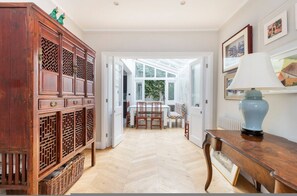 A wooden cabinet with drawers, a table with a lamp, and a view into a room with a window and a chair. - Whoops-a-daisy 3 Bedroom in Notting Hill (London)