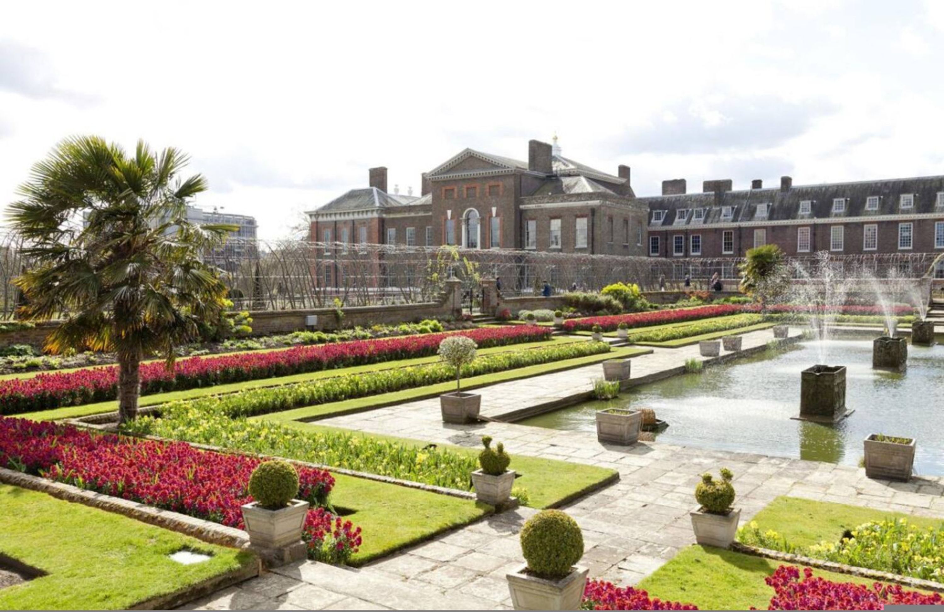 Een formele tuin met een fontein, verzorgde bloembedden en een klassiek gebouw op de achtergrond.