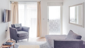 Een moderne woonkamer met een grijze bank, een grijze fauteuil, een flatscreen televisie en een groot raam met gordijnen.