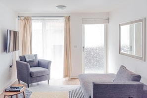 A modern living room with a grey sofa, a grey armchair, a flat-screen TV, and a large window with curtains. - The City Song 1 Bedroom Apartment in Rotherhithe (London)