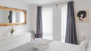 Een net opgemaakt bed met een grijze deken, een witte dressoir, een spiegel en een potplant.