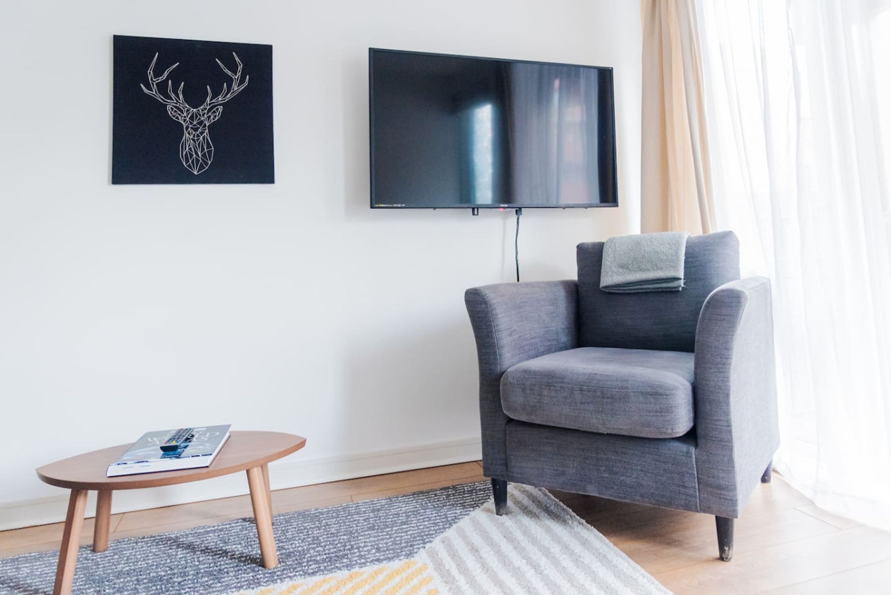 A modern living room with a grey armchair, a flat-screen TV, a framed deer head artwork, and a small wooden table with a magazine.