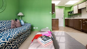 Una habitación con pared verde, un sofá estampado, una pequeña mesa con libros, una lámpara y una zona de cocina con armarios de madera y un microondas.