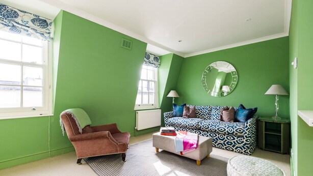 Una habitación con paredes verdes, un sillón marrón, un sofá estampado, un espejo redondo y dos lámparas.