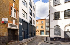 Eine schmale Straße mit Gebäuden, eines mit einem 'For Sale'-Schild, ein weiteres mit einem 'Hoxton Square'-Schild und ein drittes mit einem Wandgemälde.