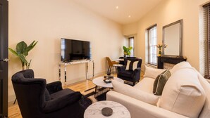 A living room with a sofa, armchair, TV, small table, and potted plants. - London Muse 1 Bedroom Apartment in Belgravia (London)