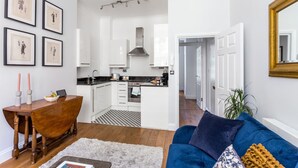Et moderne køkken med hvide skabe, en sort bordplade og en central ø. Der er et spisebord med stearinlys og en bærbar computer. Opholdsområdet har en blå sofa og et stort spejl.