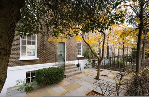 A brick building with a white door, a small porch, and a tree with yellow leaves. - London Living 3 Bedroom Home in Chelsea (London)