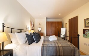 A bedroom with a bed, nightstand, lamp, wooden wardrobe, and a painting on the wall. - The Call of London 2 Bedroom Flat in Battersea (London)
