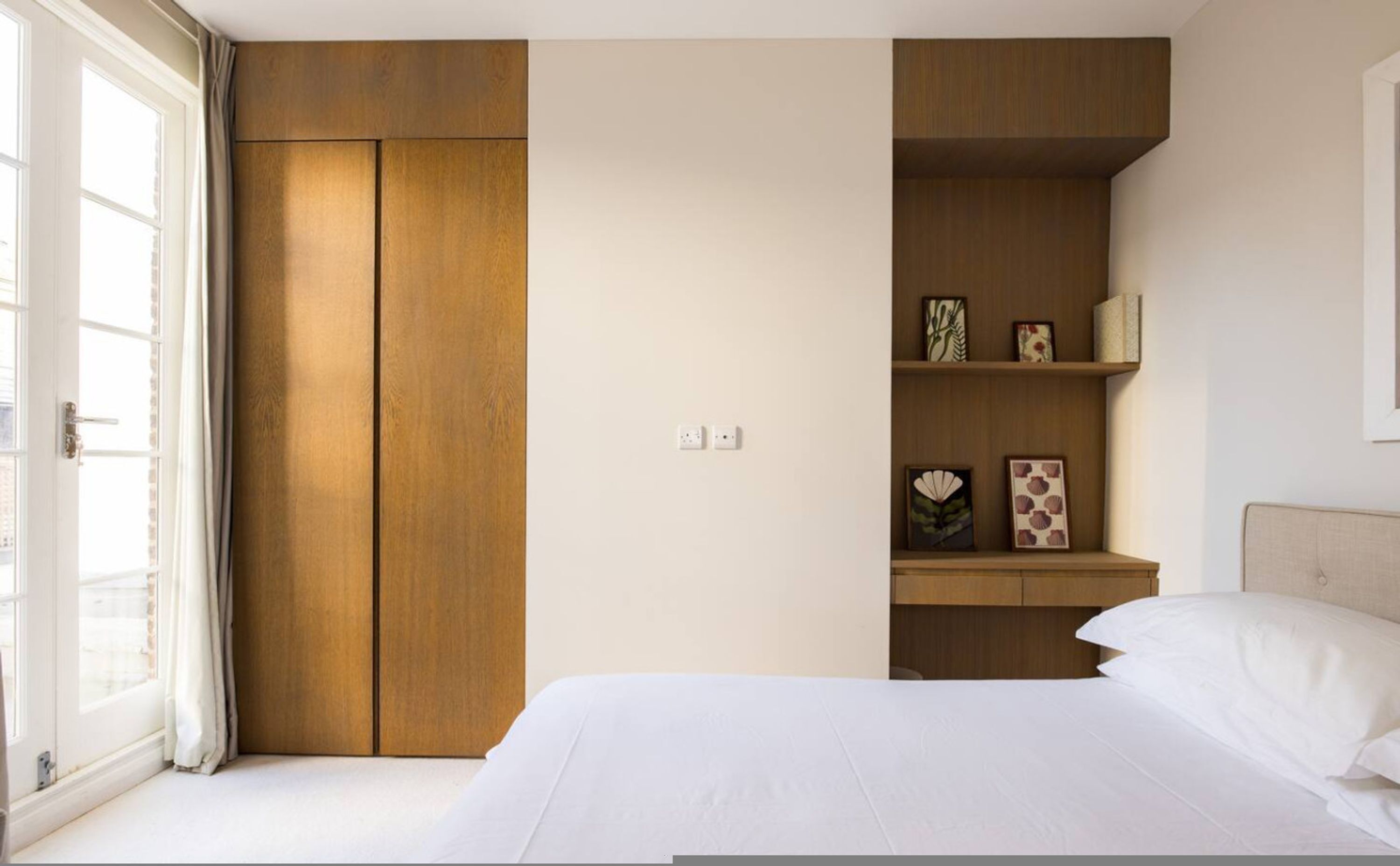 A bedroom with a bed, a wardrobe, a shelf with decorative items, and a window with curtains.