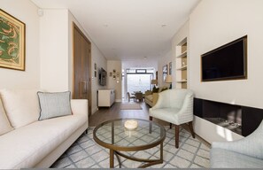 A modern living room with a sofa, armchair, glass coffee table, fireplace, and a view to the outside. - Sunny in Chelsea Luxurious 3 Bedroom Chelsea Home (London)