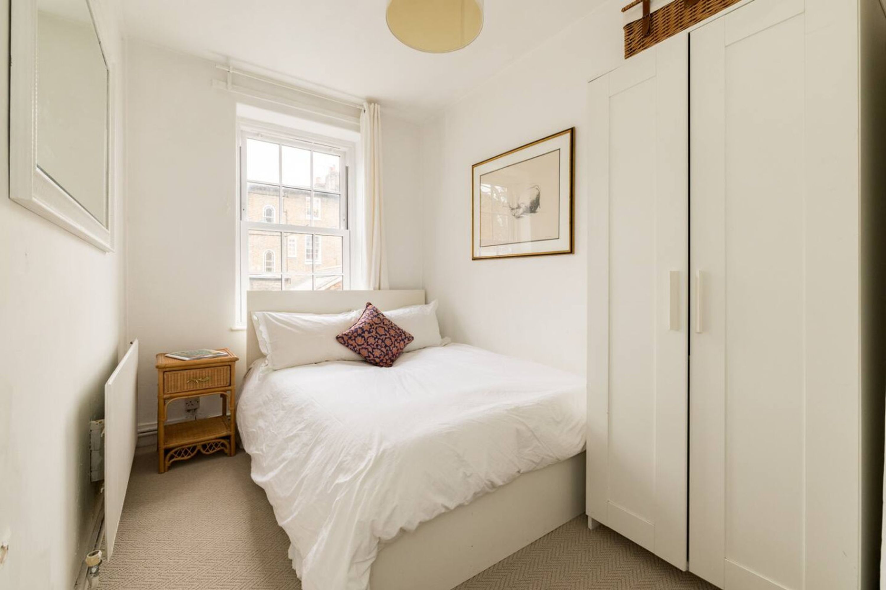 A bedroom with a bed, a window, a bedside table, a wardrobe, and a framed picture on the wall.
