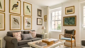 Une pièce avec un canapé gris, une chaise en bois, une table basse blanche et, au mur, des illustrations d’oiseaux encadrées.