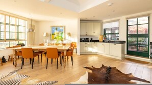 A spacious kitchen with white cabinets, a dining area with orange chairs, and a large window with a view of trees. - A Gentle Riverside Charm 2 Bedroom Mews House (London)