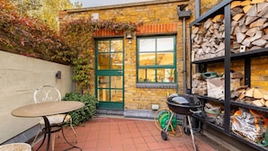 A brick building with a green door, a small patio with a table and chairs, and a stack of firewood. - A Gentle Riverside Charm 2 Bedroom Mews House (London)