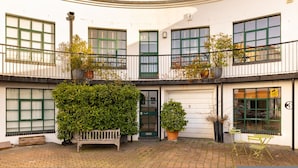 A building with a curved facade, multiple windows with green shutters, a bench, and potted plants. - A Gentle Riverside Charm 2 Bedroom Mews House (London)