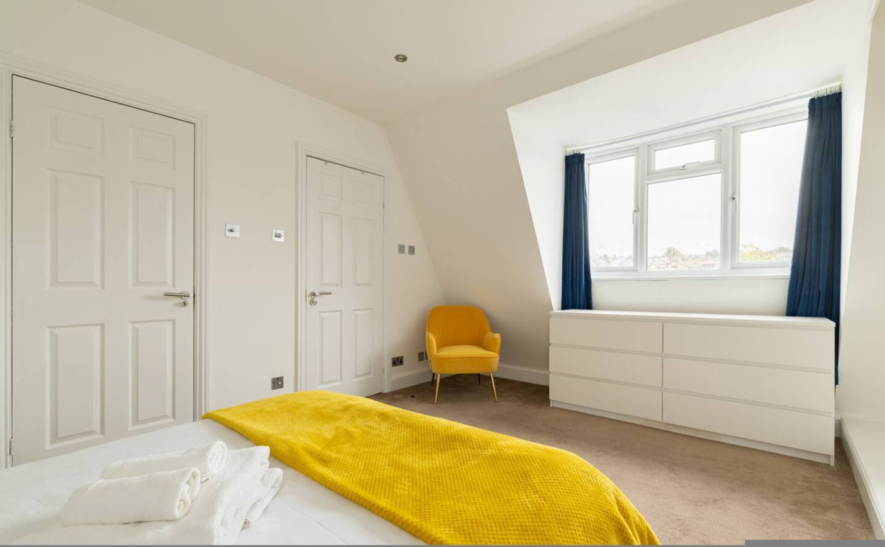 A bedroom with a bed, a yellow chair, a window with curtains, and white wardrobes.