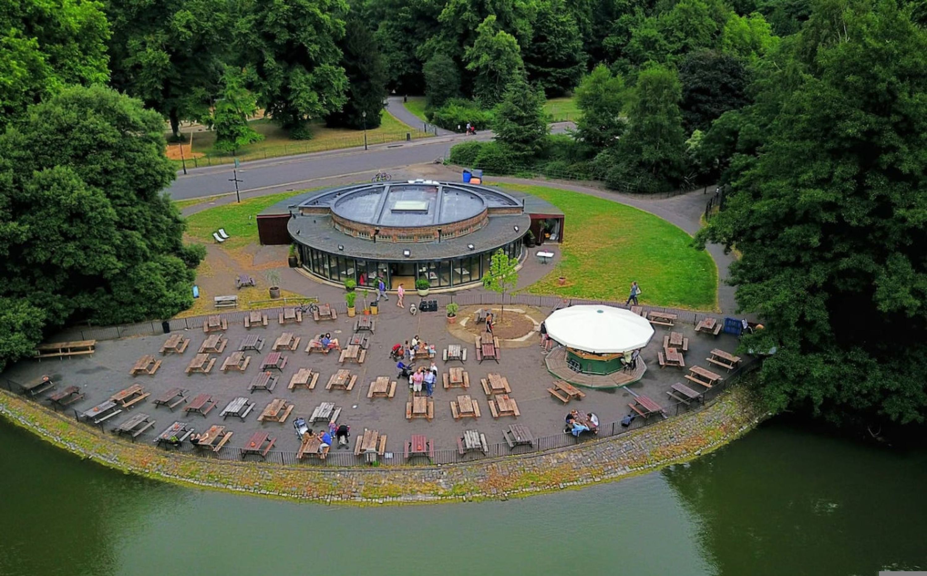 A circular outdoor area with wooden benches and a central circular structure, surrounded by trees and a body of water.