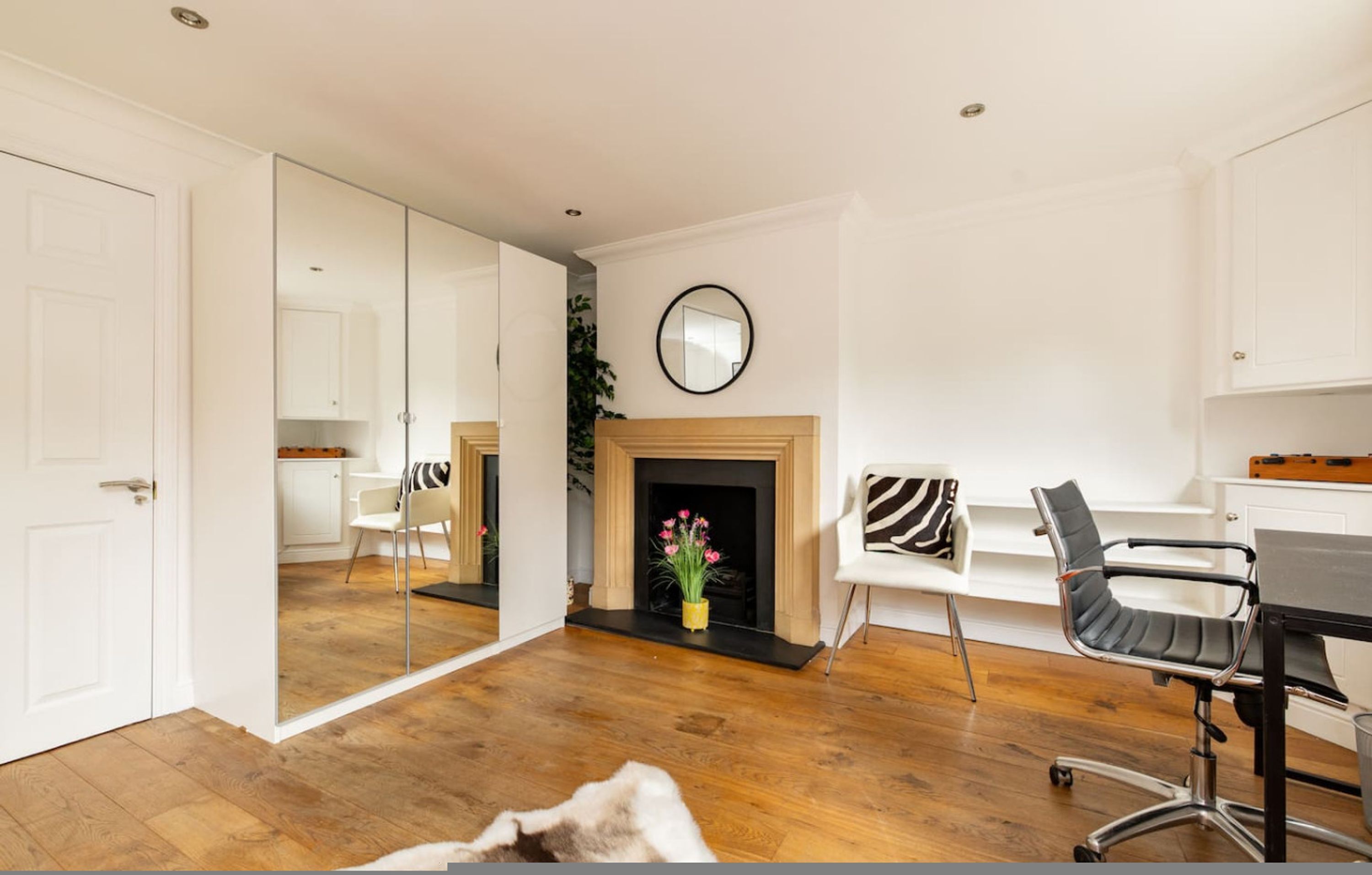 A room with a wooden floor, a fireplace with pink flowers, a mirror on the wall, and a dining area with a table and chairs.