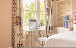 A bedroom with a bed, a vanity with a mirror, a stool, a lamp, and a window with floral curtains. - Battersea Park 3 Bedroom House With Private Garden (London)