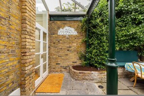 A brick wall with a door, a sign with a tree and the word 'STANLEY'S,' a green bench, and a small garden bed. - Battersea Park 3 Bedroom House With Private Garden (London)