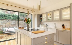 A modern kitchen with a large island, built-in appliances, and a view of a garden. - Your Zen - 2 Bedroom Mews House in Notting Hill (London)