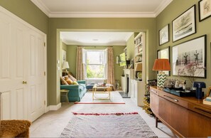 Ein Wohnzimmer mit Sofa, Couchtisch und Kamin. Links befindet sich eine Tür und rechts ein Bücherregal.